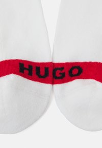 Weiße Socken mit einem dicken roten Band, auf dem das Wort "HUGO" in schwarzer Schrift steht. Der Stoff wirkt glatt und hat ein enganliegendes, sportliches Design.