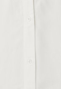 Gros plan sur le devant d'une chemise blanche montrant la texture du tissu et deux boutons blancs translucides sur la patte de boutonnage.