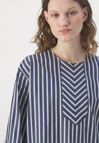 Chemise rayée bleu et blanc avec un col rond, présentant un détail en chevrons sur la patte de boutonnage et des manches amples. Texture de tissu lisse.