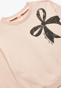 Jasnoróżowy sweatshirt z okrągłym dekoltem z dużym czarnym wzorem graficznym przedstawiającym wstążkę oraz tekst na przodzie. Miękka, gładka faktura.