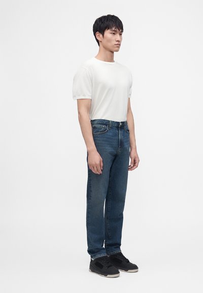 Jeune homme debout portant un t-shirt blanc uni, un jean bleu et des baskets noires sur un fond blanc.