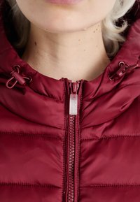 Bordo puffer jakna s visokim ovratnikom. Ima središnji patentni zatvarač, podesive vezice i kviltanu teksturu.