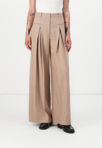Pantaloni a gamba larga beige a righe con pieghe frontali, passanti per cintura e tasche laterali, abbinati a scarpe nere basse. Tessuto di consistenza liscia.