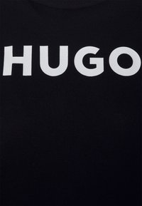 Mustaa kangasta, jossa keskellä on suuret valkoiset kirjaimet, jotka lukevat "HUGO".