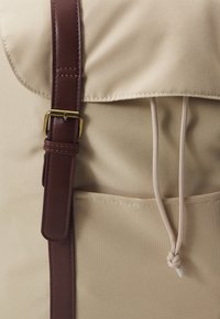 Sac à dos en tissu beige avec une sangle en cuir marron et une boucle en laiton, doté d'une fermeture à cordon, complété par une poche avant.