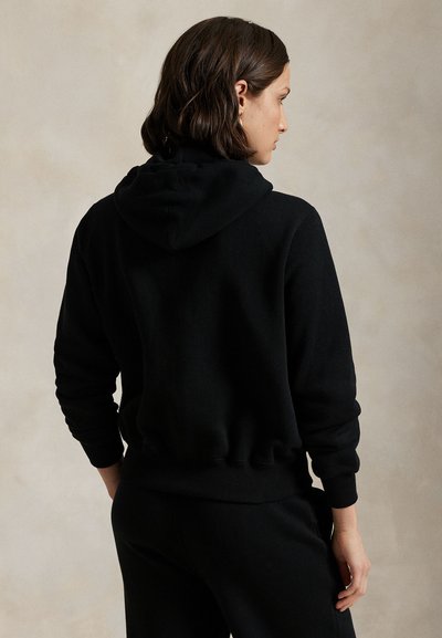 Osoba s krátkými tmavými vlasy, oblečená v černé mikině s kapucí a černých kalhotách, stojící zády k neutrálnímu pozadí.