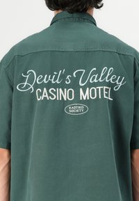Zöld, rövid ujjú ing, amelyen fehér hímzett felirat található: "Devil's Valley CASINO MOTEL" és "KAOTIKO SOCIETY" embléma a hátsó oldalon. Pamut anyagból készült.