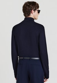 Marinefarbener Rollkragenpullover mit langen Ärmeln, feiner Strickstruktur und schmaler Passform. Kombiniert mit marineblauen Hosen und einem schwarzen Leder-Gürtel.