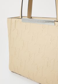 Beige tote bag laget av teksturert materiale, med et gjentakende logo-mønster, svarte detaljer og sølvfargede håndtak.