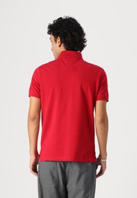 Rød polo-shirt med krave og korte ærmer. Glat tekstur. Tilpasset pasform med sideslids ved kanten. Grå bukser synlige nedenfor.