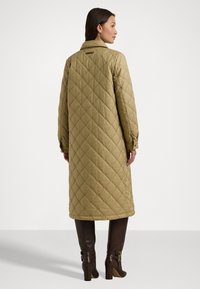 Alyvuogių žalumos siuvinėta ilga striukė su apykakle, mygtukais užsegamomis rankovėmis ir tekstūruotu rombų raštu. Suderinta su tamsiai ruda iki kelių siekiančiomis botais.