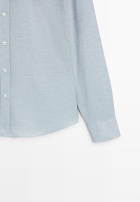 Camicia azzurro chiaro con tessuto strutturato, orlo sagomato e maniche lunghe, con bottoni bianchi sulla parte frontale.