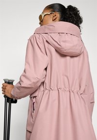 Lys rosa vanntett jakke med hette, innsvingt midje, frontlomme med glidelås og elastiske mansjetter. Tilbehørt med turgåerpinner.