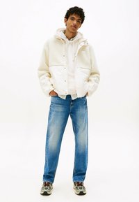 Weißer, flauschiger Stoffjacke mit einem glatten unteren Abschnitt, kombiniert mit hellblauen gerade geschnittenen Jeans und grauen Sneakers mit dunklen Akzenten.