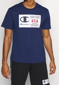 Muž v námořnickém tričku Champion s velkým logem na přední straně a červeným textem "U.S.A.", doplněný černými šortkami se shodným nášivkou.