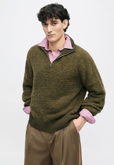 Olivgrüner Fleece-Pullover mit halbem Reißverschluss und Kragen, kombiniert mit einem hellrosa Hemd mit Kragen. Rippstrick an den Bündchen und dem Saum. Braune Hose.