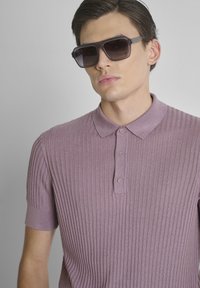 Joven con gafas de sol negras grandes y una camisa polo de canalé de manga corta color malva, posando frente a un fondo gris liso.