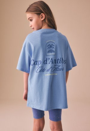 Ung pige med langt hår iført en oversized lyseblå t-shirt og blå shorts, står sidelæns mod en beige baggrund.