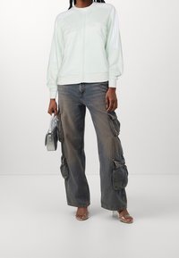 Mintově zelená bunda na zip s bílými pruhy, spárovaná s vybledlými cargo džíny se bočními kapsami a průhlednými sandály na podpatku.