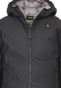 Giubbotto imbottito nero con chiusura a zip, dotato di una fodera interna grigia e un patch con logo sul petto. Texture esterna liscia, design con cappuccio.