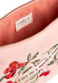 Vaaleanpunainen kangaspussi, jossa on kukkakirjontaa ja kultainen vetoketju. Sisällä on ommeltu etiketti, jossa lukee "FABLE ENGLAND". Pehmeä rakenne.