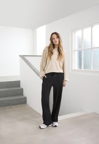 Cream long-sleeve top with pleated sleeves, paired with black loose-fitting pants and gray sneakers; set against a minimalist interior.