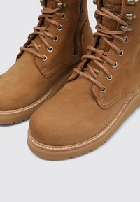 Botas de tobillo de cuero marrón con puntera redonda, acentos metálicos y diseño de cordones. Presentan una suela texturizada contrastante y detalles de costura.