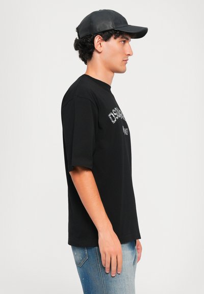 Giovane uomo in piedi di lato, con berretto di pelle nera, t-shirt nera oversize e jeans blu sbiaditi, su uno sfondo bianco semplice.