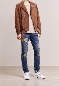 Ruskea mokkanahkayksi pyöräilijän takki, jossa on vetoketjulliset taskut, yhdistettynä revittyihin sinisiin farkkuihin ja valkoisiin lenkkareihin.