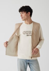 Beige gesteppte Weste mit Reißverschluss, offen über einem cremfarbenen Sweatshirt getragen. Verfügt über ein dreieckiges Logo und hat eine glatte, leichte Textur.