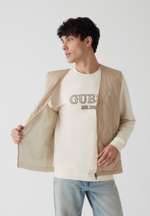 Chaleco acolchado beige con cierre de cremallera, mostrado abierto sobre un sudadera color crema. Presenta un logo triangular y tiene una textura suave y ligera.
