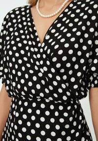 Schwarzes Wickelkleid mit großen weißen Punkten, kurzen Ärmeln und tailliertem Schnitt. Verfügt über einen V-Ausschnitt und eine glatte Stofftextur.