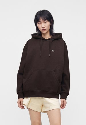Jeune personne aux cheveux noirs portant un sweat à capuche Adidas marron foncé et un short clair sur un fond blanc uni.