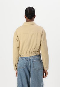 Henkilö, jolla on letitetty hiuskuontalo matalassa nutturassa, pukeutuneena beigeen takkiin, jossa on joustava vyötärö ja sinisiin korkeavyötäröisiin farkkuihin, nähtynä takaa.