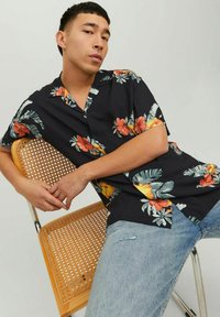 Camicia nera a maniche corte con motivo floreale presenta fiori rossi e gialli vivaci con foglie verdi, texture simile al cotone. Il modello indossa jeans in denim.