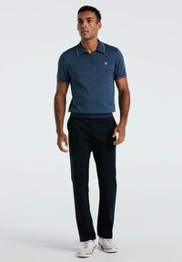 Homme debout portant un polo à manches courtes bleu à motifs, un pantalon noir et des baskets blanches sur un fond gris clair uni.