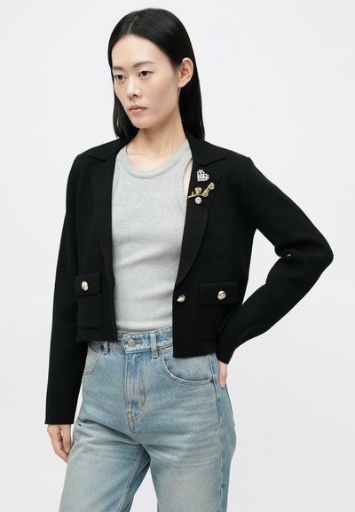 Μαύρο cropped σακάκι με κοφτό πέτο, μπροστινές τσέπες, χρυσά κουμπιά και διακοσμητικά μπροσάκια. Φοριέται πάνω από ανοιχτό γκρι τοπ και μπλε τζιν.