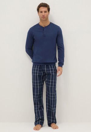 Hombre de pie descalzo vistiendo una camiseta henley de manga larga azul marino y pantalones de pijama de cuadros azul marino frente a una pared lisa.