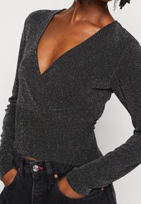 Svart långärmad topp med djup V-ringning, tillverkad av ett glittrande tyg med texturerad yta, kombinerad med mörka jeans.