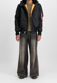 Veste bombardier noire avec doublure orange, associée à un jeans sombre, large et délavé. Caractéristiques : poignets côtelés et jambes de pantalon larges.