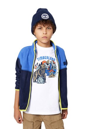 Jachetă din polar albastră și bleumarin cu accente verde neon, purtată peste un tricou alb cu grafică Timberland, care prezintă un urs ilustrat și munți.