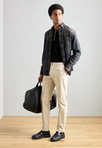 Denim shirt in dark blue, black polo, beige pants, black shoes, and a black duffle bag with side seams. Natural wood floor and light background.