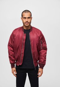 Veste aviateur bordeaux avec fermeture éclair, ourlet et poignets côtelés, poches latérales et poche à fermeture éclair sur la manche, portée par-dessus une chemise noire.