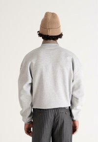 Felpa grigia con spalle scese, colletto a coste e un'etichetta sull'orlo, abbinata a pantaloni a righe grigie e nere e a un berretto lavorato a maglia beige.