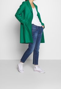 Abrigo de lana verde con solapa a medida, combinado con una camiseta blanca, jeans azules y zapatillas blancas de caña alta. Diseño simple y estructurado.
