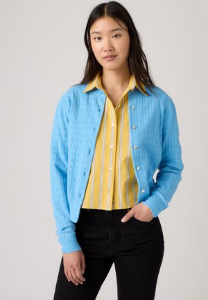 Jeune femme portant un cardigan bleu boutonné sur une chemise jaune rayée avec un pantalon noir, debout avec une main dans la poche.