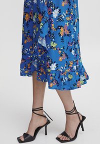 Blå blommig klänning med kort volangkant, med gula, vita och rosa blommor. Svarta högklackade sandaler med knytband runt ankeln.