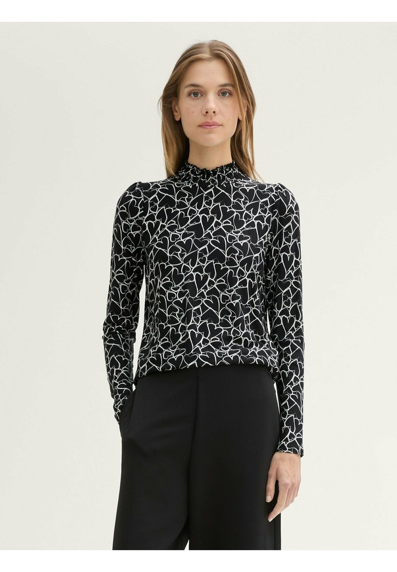 Zwarte top met lange mouwen en een wit hartpatroon, met een hoge, geraffineerde halslijn en een gladde textuur. Gecombineerd met zwarte broeken.