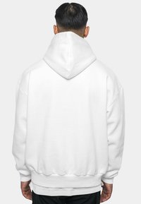 Haut à capuche blanc oversized avec une capuche, des poignets et un ourlet côtelés. Tissu doux au toucher lisse, sans motifs ni accents visibles.