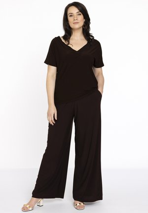 Zwarte V-hals top met korte mouwen, gecombineerd met losse, wijde zwarte broek. Het model draagt witte blokhakken sandalen.